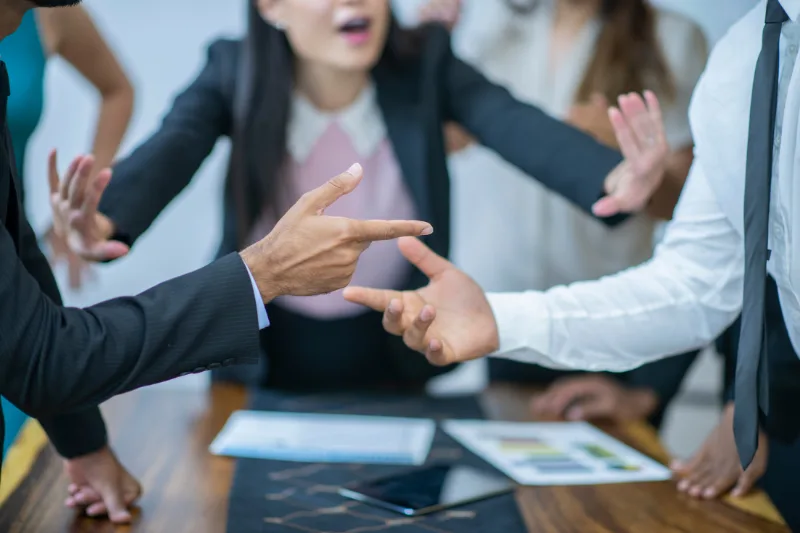 Varias personas en una discusión acalorada, señalándose con las manos en una mesa de oficina.