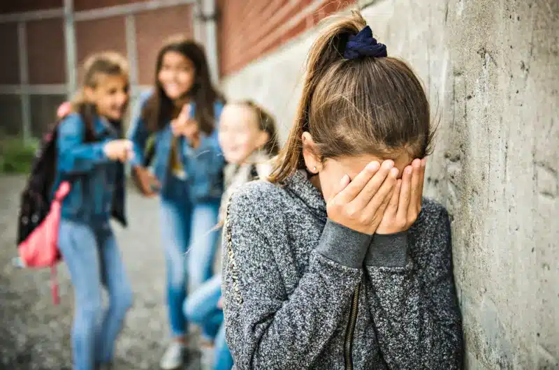 Niña con las manos en la cara sufriendo acoso escolar mientras otras compañeras se ríen y señalan